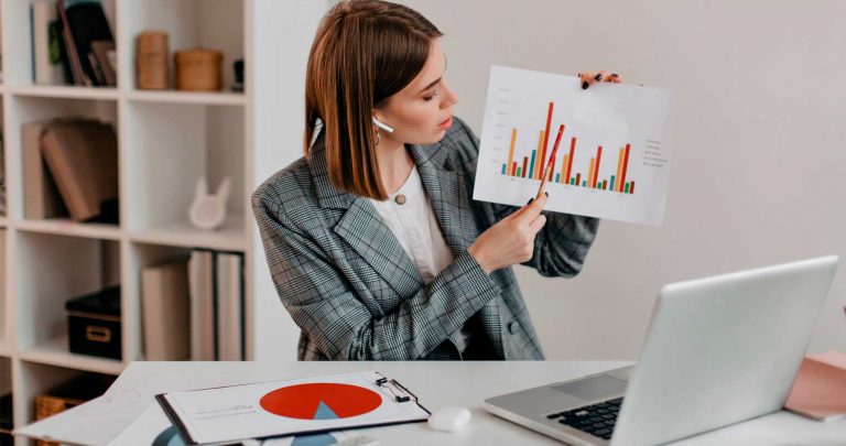 Uma executiva sentada na frente de um notebook com um gráfico na mesa e outro na mão, mostrando indicadores de produtividade.