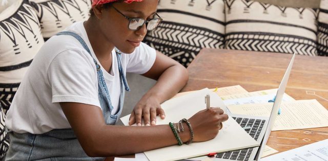 mulher negra, de óculos, sentada em uma mesa com notebook e papeis, trabalhando na criação de conteúdo