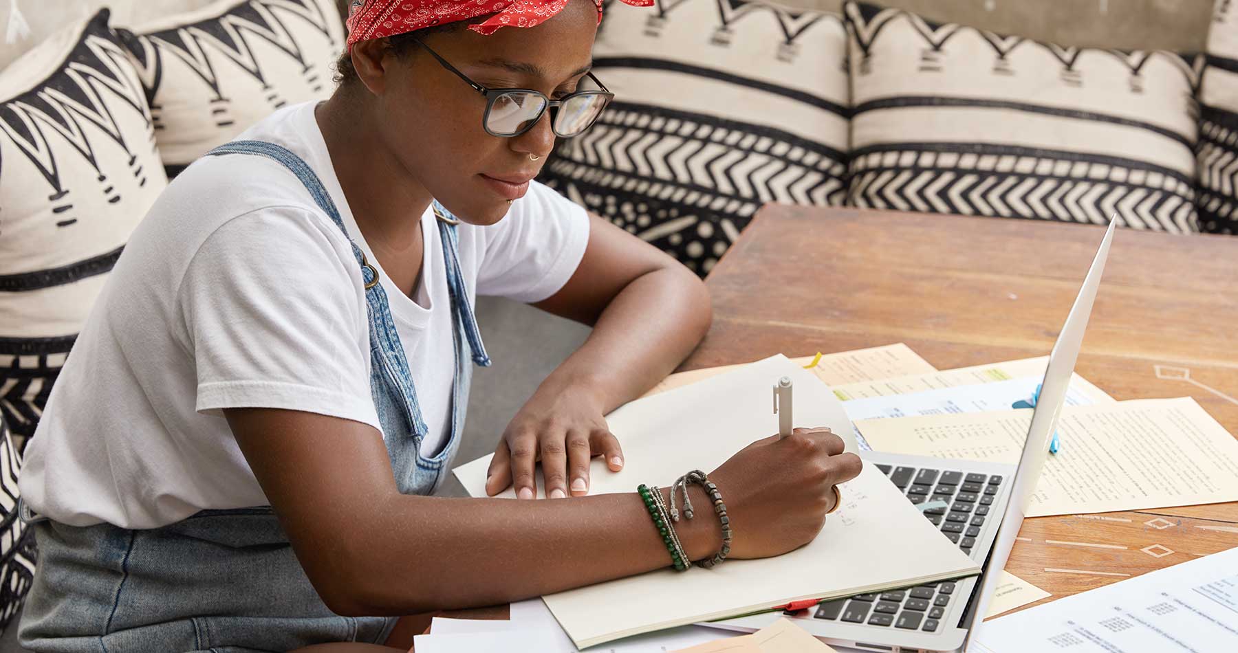 mulher negra, de óculos, sentada em uma mesa com notebook e papeis, trabalhando na criação de conteúdo