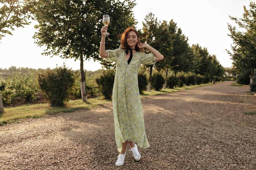 Jovem com vestido floral e tênis passeando no campo com uma taça de vinho na mão na tendência Athleisure