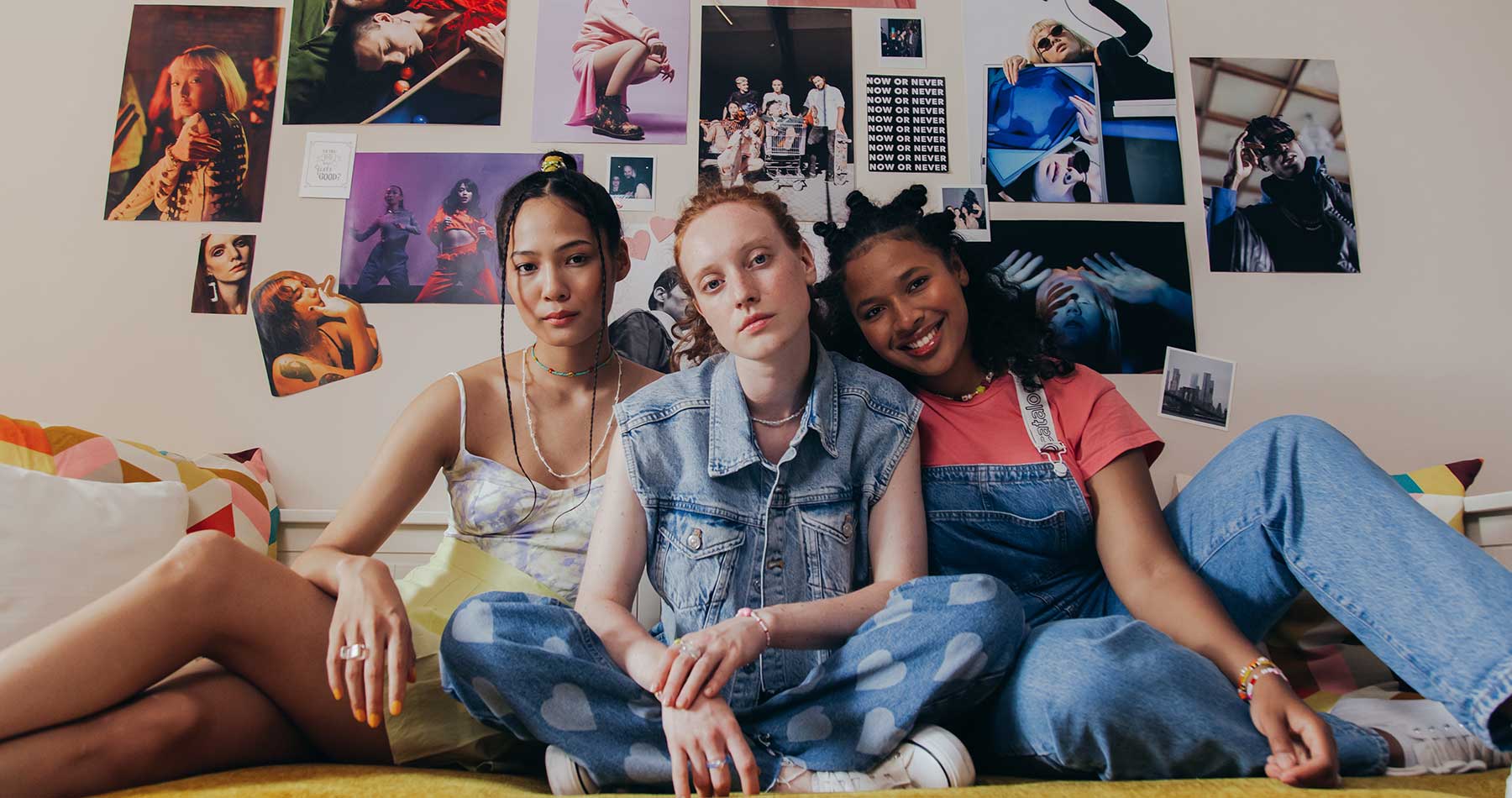 foto de três meninas, sentadas confortavelmente sobre um colchão, em frente a uma parede com diversos pôsteres em referência a tendência anos 80