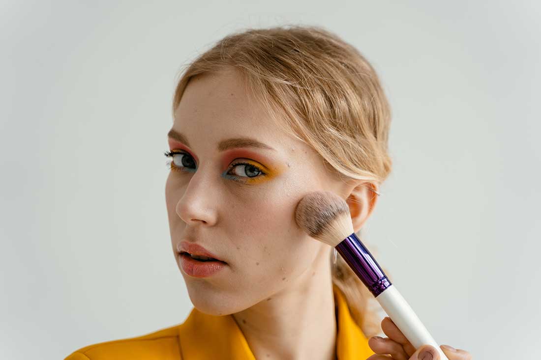 foto de mulher loira com o cabelo preso, aplicando maquiagem, nos olhos a maquiagem tem cores vibrantes, com tons de laranja, azul e rosa