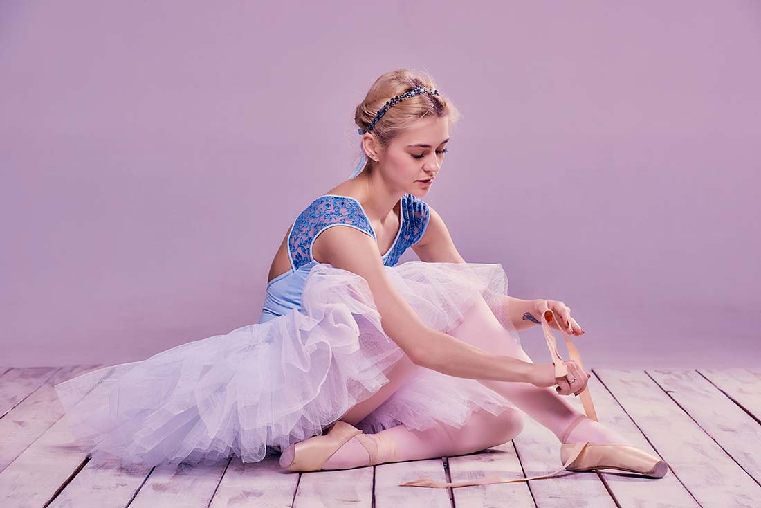 imagem de bailarina sentada no chão, mexendo em sua sapatilha, que possui amarração de fita de cetim, para demonstrar uma das possibilidades de aplicação das fitas rígidas no vestuário