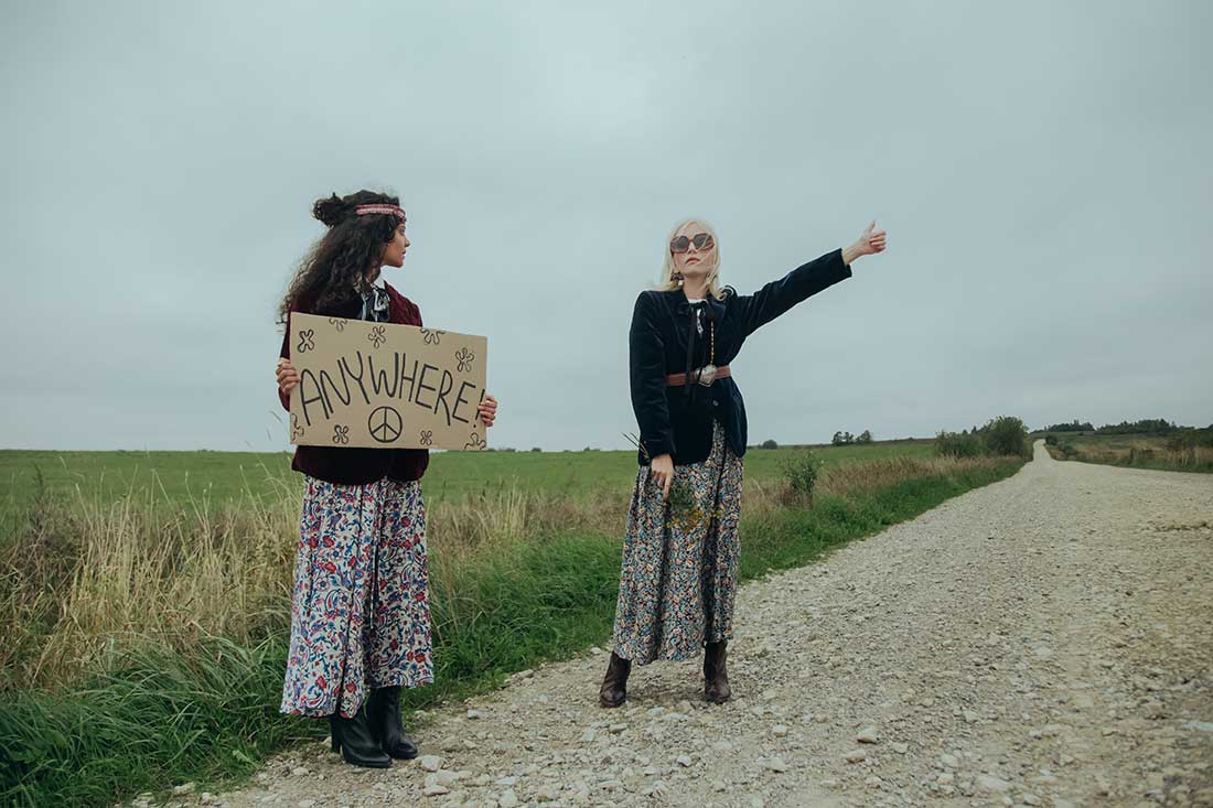 duas mulheres vestidas de acordo com o estilo hippie, pedindo carona em uma estrada, segurando uma placa