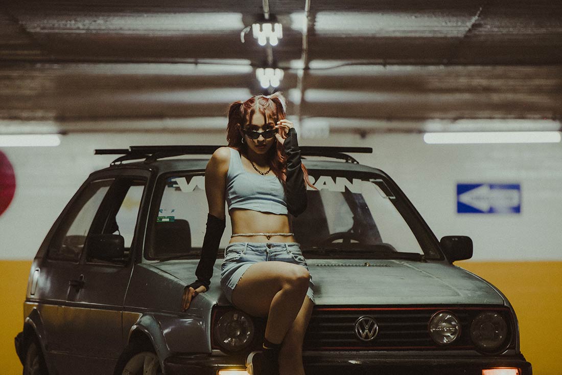 mulher de cabelo preso e óculos escuros posando para foto sobre o capô de um carro, usando luvas